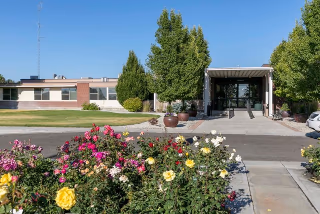 Front entrance of a single-story care facility with a covered walkway, trees, and colorful rose beds in the foreground.