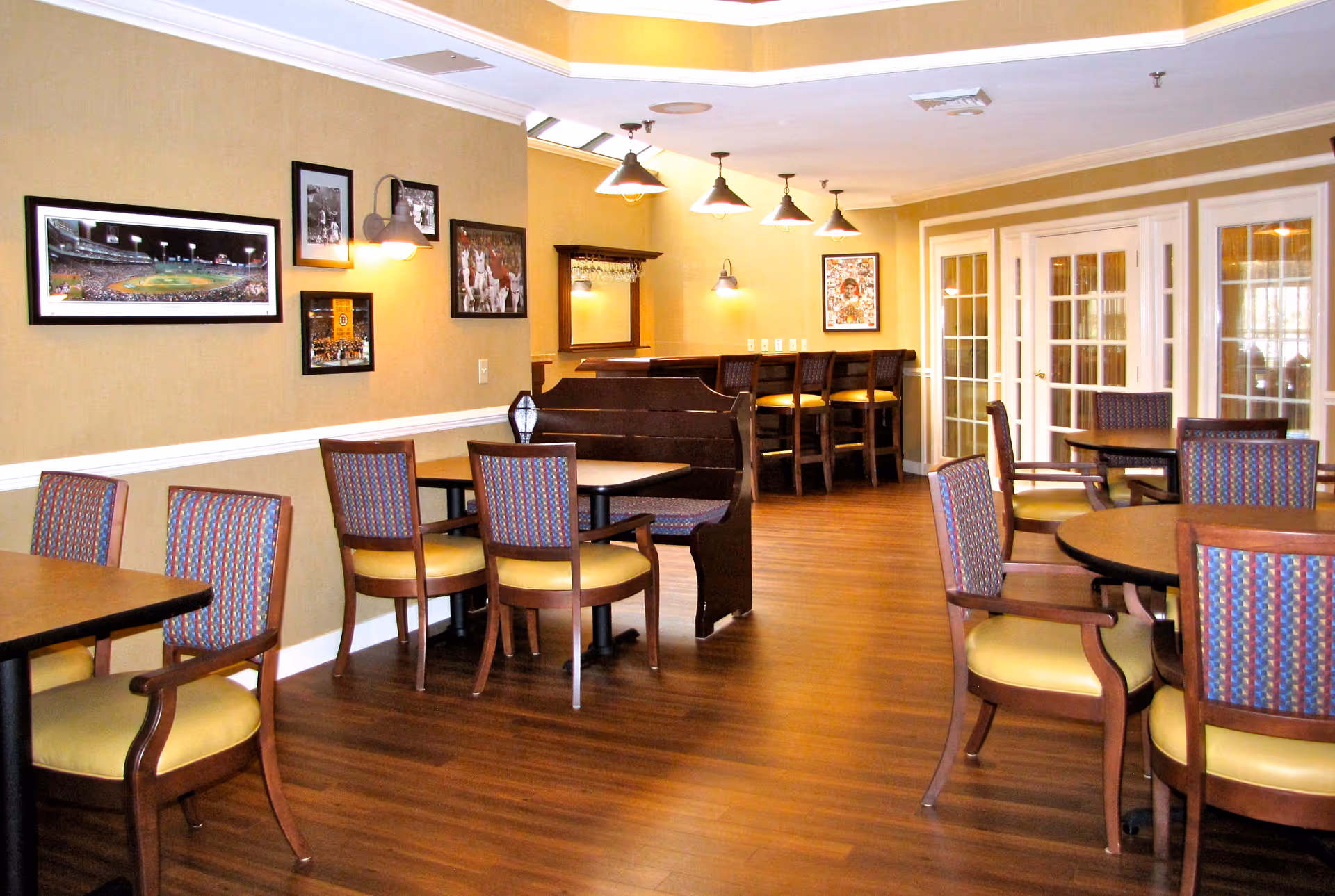 A well-lit dining area with wooden floors, multiple tables and chairs with patterned upholstery, a booth seating area, framed sports-themed pictures on the walls, and pendant lights hanging from the ceiling. French doors are visible in the background.