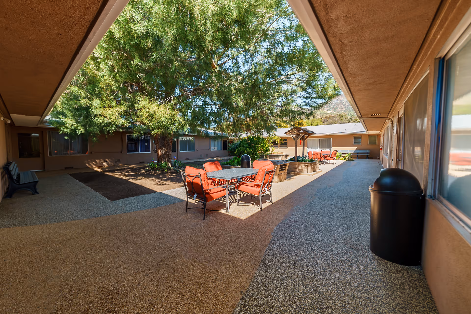Outdoor courtyard area with a large tree in the center, surrounded by a building with windows. There are several tables with red cushioned chairs and benches along the walls. The area is well-lit with natural sunlight.