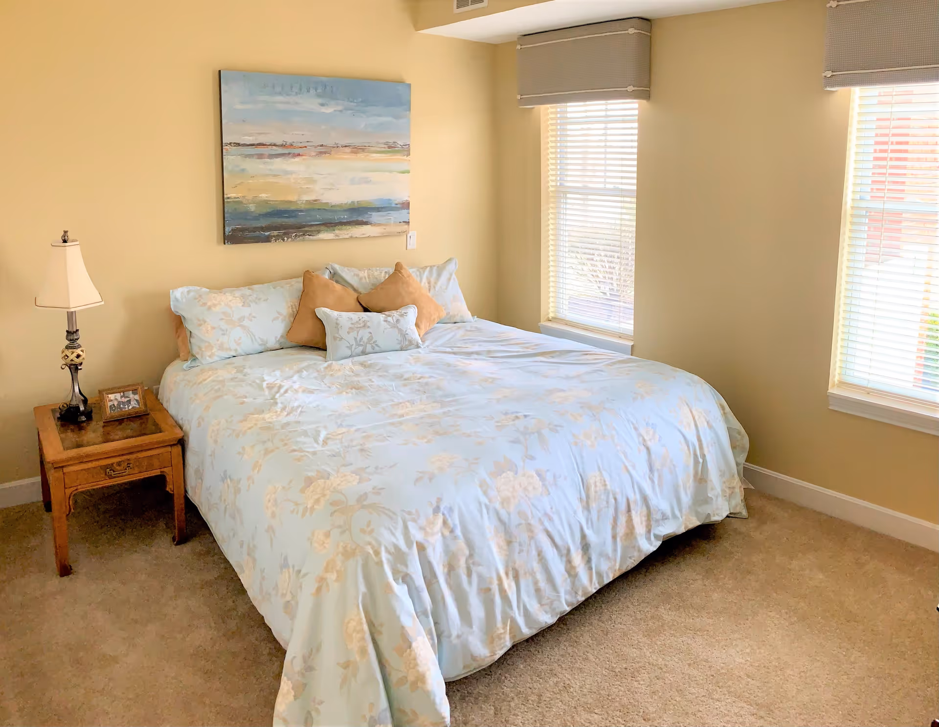 A bedroom with a large bed covered in light blue floral bedding and several pillows. There is a wooden nightstand with a lamp and a framed photo on it. Two windows with blinds allow natural light into the room. A colorful abstract painting hangs on the beige wall above the bed.