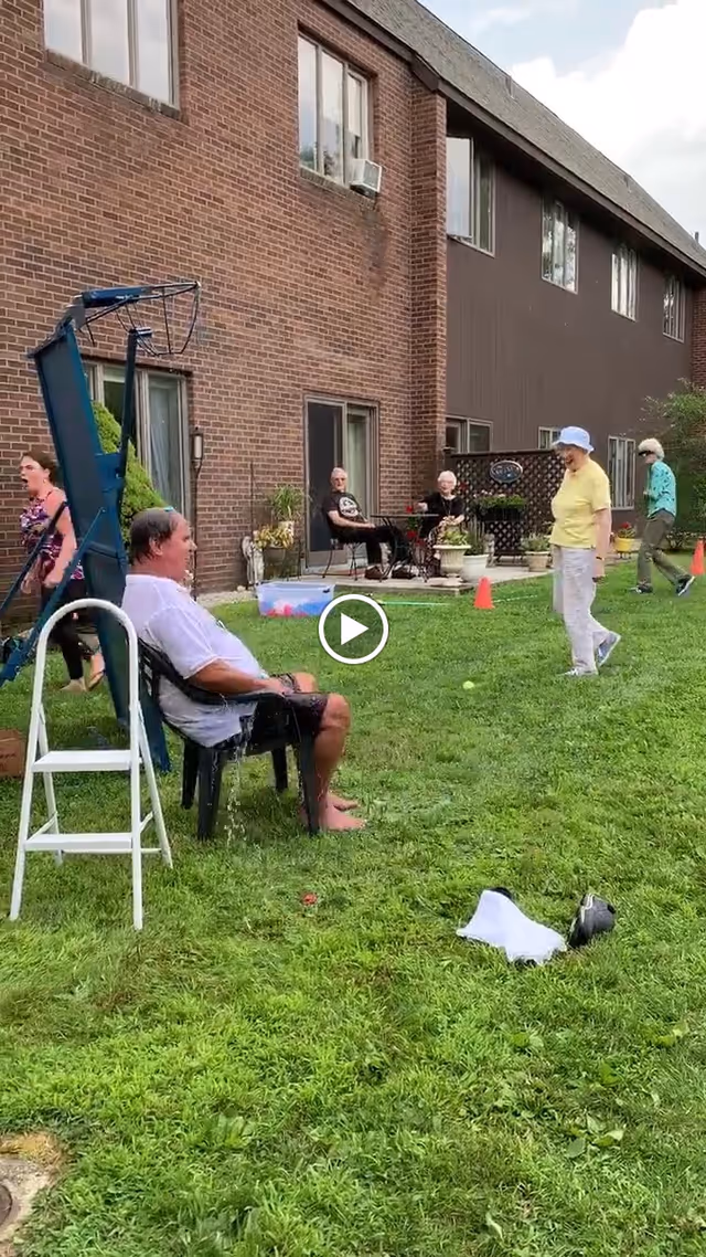 Several elderly people are outside on a grassy area next to a brick and brown building. One man is sitting on a chair under a basketball hoop, appearing wet, while others stand or sit nearby. There are some orange cones and a plastic container on the grass, suggesting an outdoor activity or event.