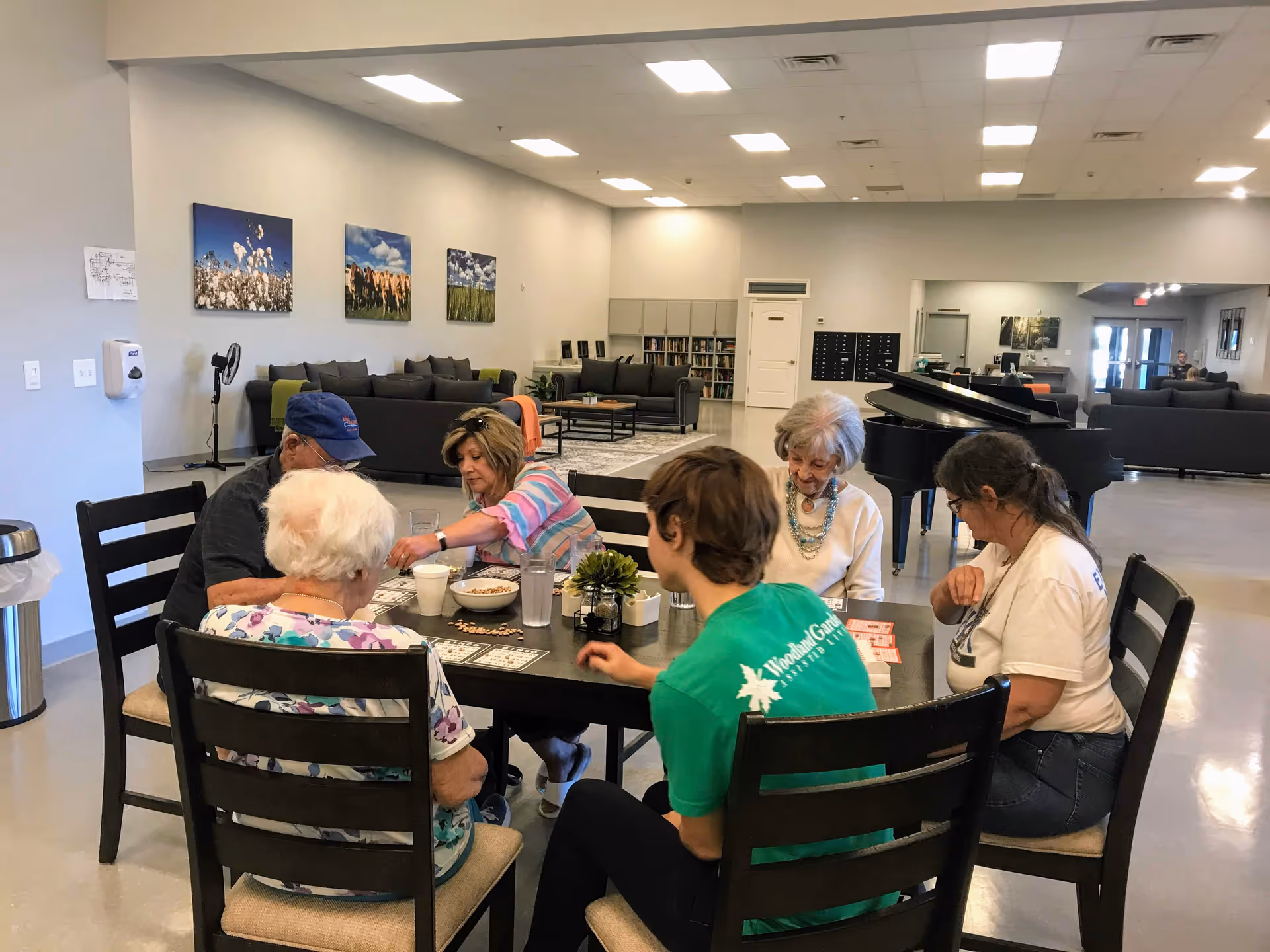 A group of six people sitting around a table playing a game in a spacious, well-lit common area with couches, a grand piano, and artwork on the walls in the background.