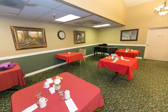 A dining room with several tables covered in red tablecloths, each set with white napkins, cups, and small flower arrangements. The room has green patterned carpet, beige walls with green wainscoting, framed paintings, a clock on the wall, and a black grand piano in the corner.