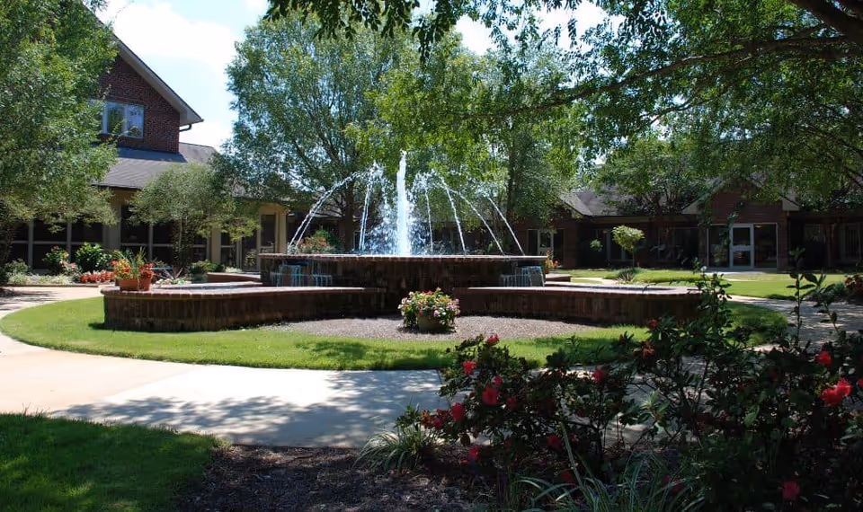 A landscaped courtyard with a multi-tier brick fountain surrounded by trees, flower beds, and low brick buildings.