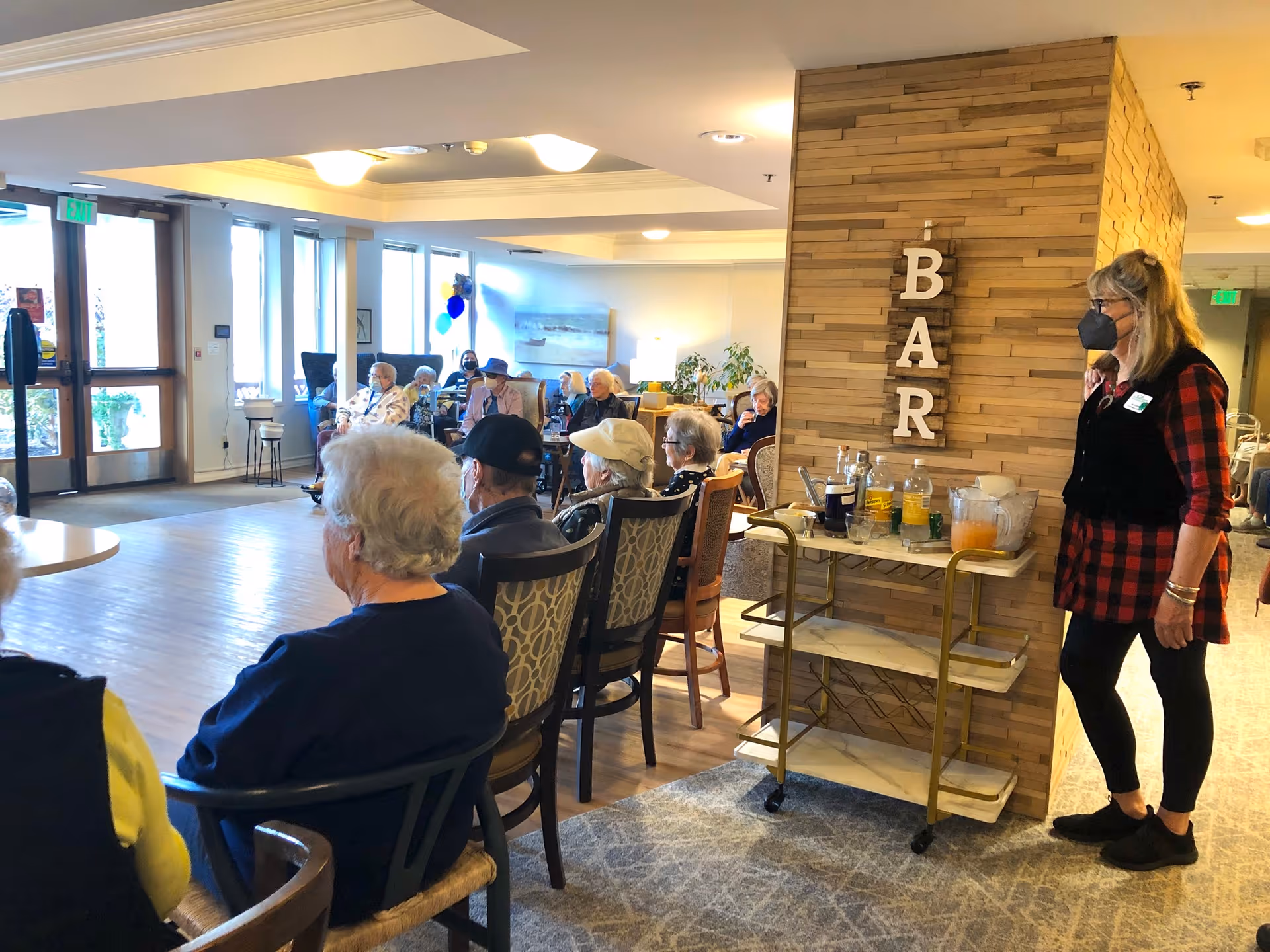 A group of elderly people seated in chairs facing a common area in a senior living facility. A woman wearing a mask and a red plaid shirt stands near a small bar cart with drinks and glasses. The wall behind the bar cart has wooden paneling with the word 'BAR' displayed vertically. Large windows and glass doors are visible in the background, letting in natural light.