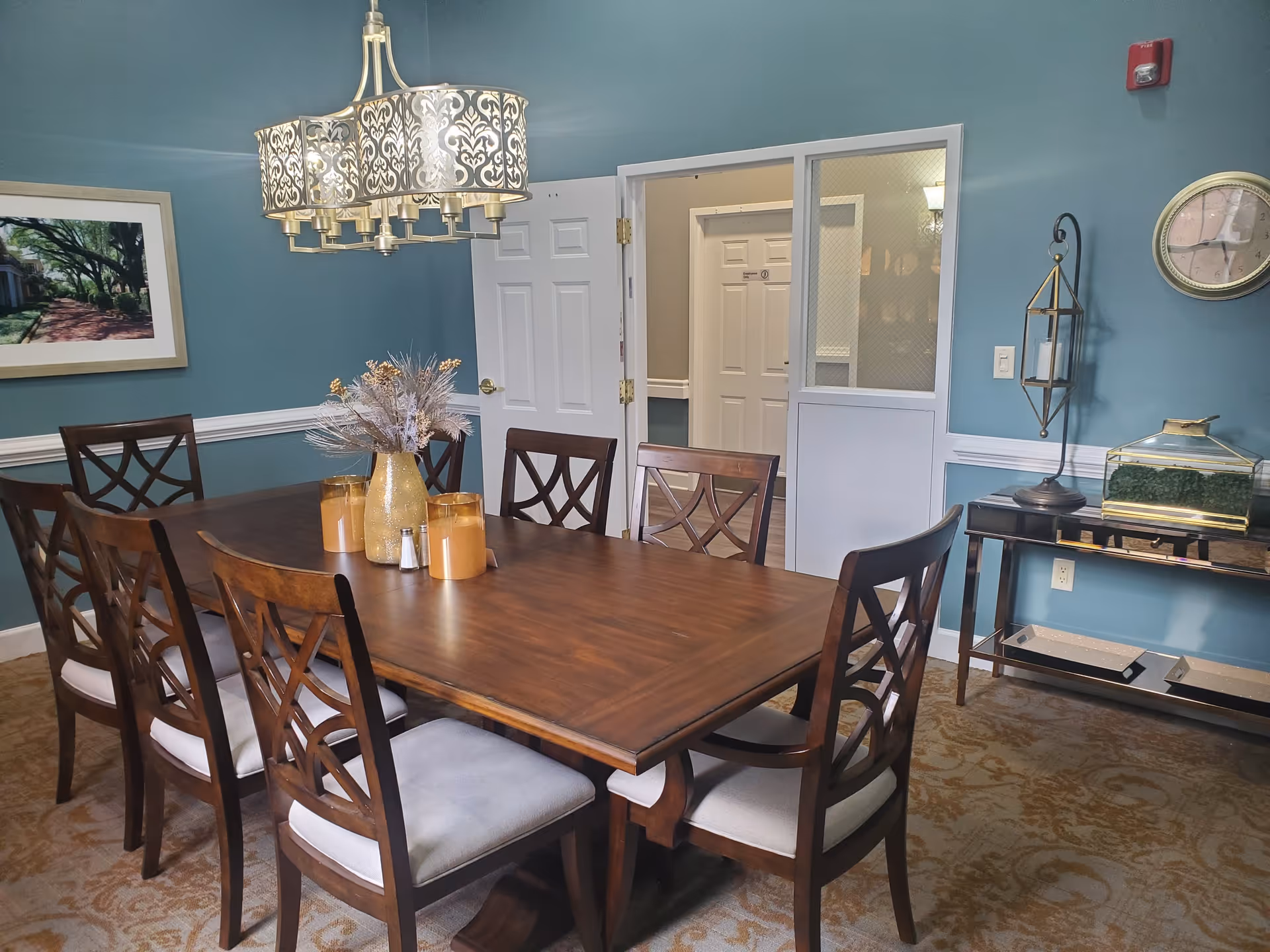 A dining room with a large wooden table surrounded by eight cushioned chairs. The table has a centerpiece with a vase and decorative items. The walls are painted teal with white trim, and there is a decorative chandelier hanging above the table. A framed picture is on the left wall, and a clock and candle holder are on the right wall. There is a doorway leading to another room with white doors.