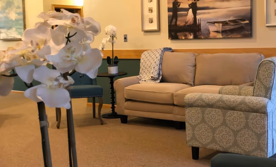 A cozy living room area with a beige sofa, a patterned armchair, a small round side table with a white orchid plant, and a blue chair. The walls are decorated with framed artwork, including a painting of a couple by a boat on the water.