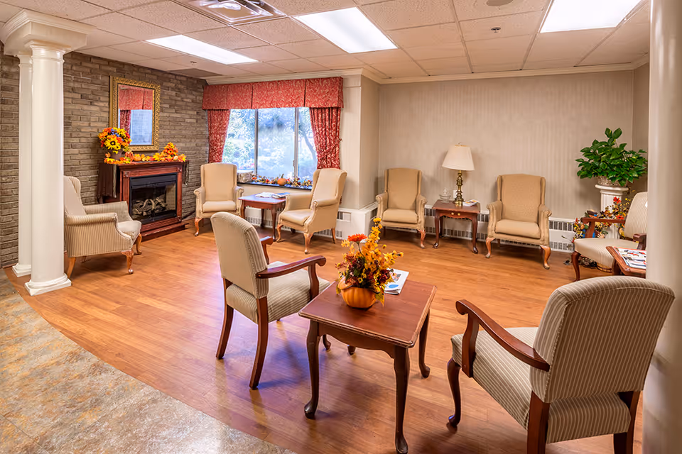 A cozy sitting area with several upholstered armchairs arranged around wooden tables. The room features a faux fireplace decorated with autumn-themed flowers and a mirror above it. There is a large window with red curtains letting in natural light, a floor lamp, and a potted plant in the corner. The floor is a combination of wood and tile, and the ceiling has recessed lighting.
