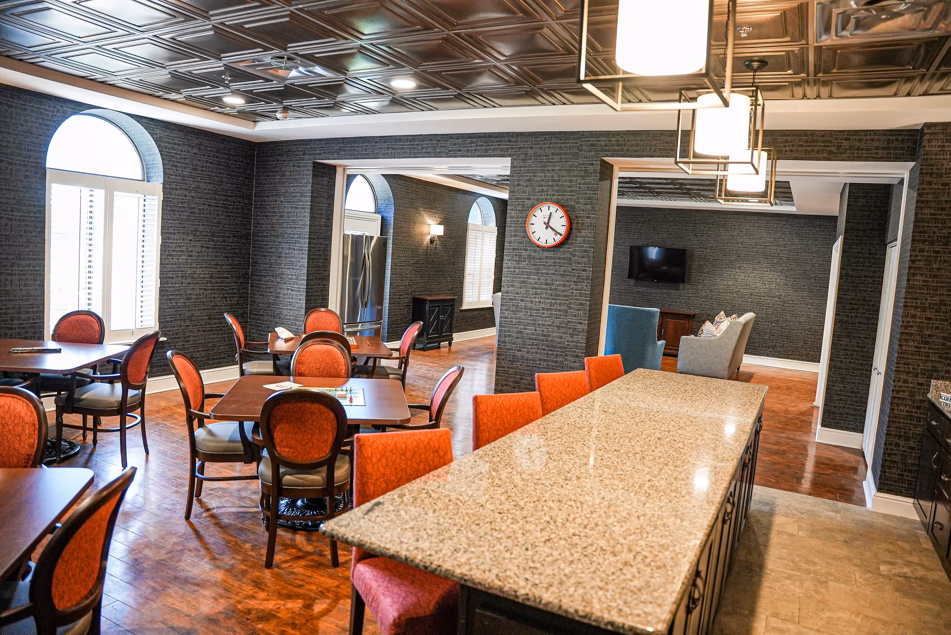 A spacious senior living facility common area with multiple tables and chairs featuring red upholstery. The room has large arched windows with white shutters, dark textured walls, and a decorative ceiling. A long granite countertop with red chairs is in the foreground, and a seating area with a TV is visible in the background.