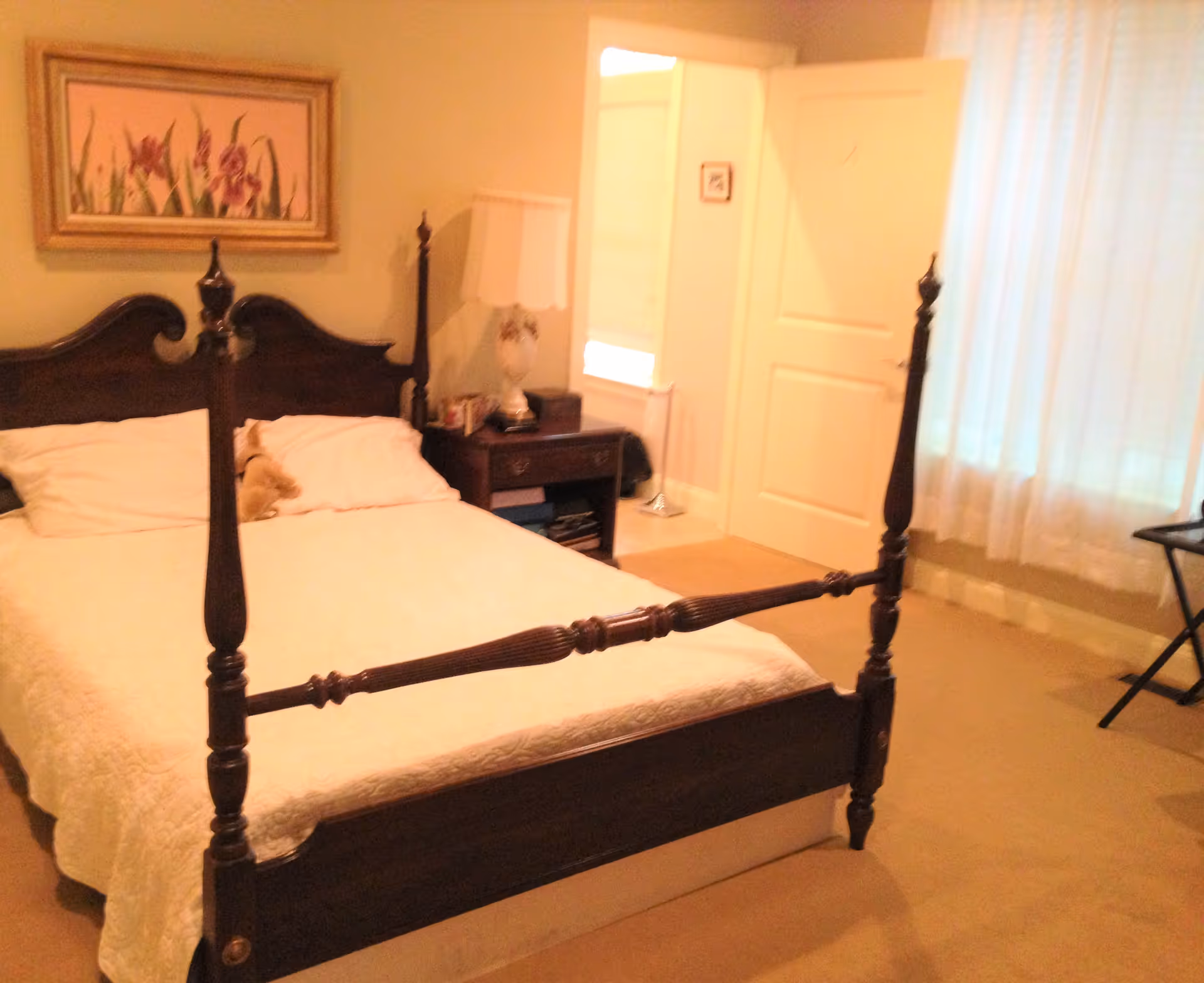 Cozy bedroom with a wooden four-poster bed, nightstand and lamp, and a doorway with a curtained window.
