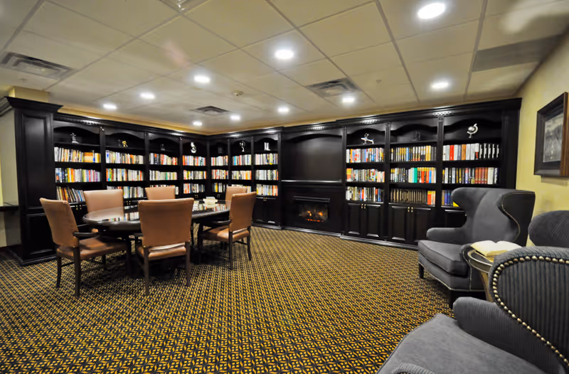 Cozy communal library-style lounge with dark wood built-in bookshelves, a fireplace, a round table with chairs, and upholstered armchairs.