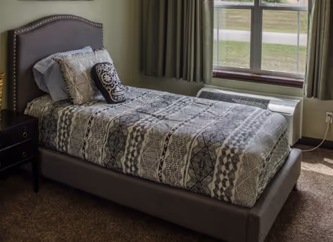 A neatly made single bed with a patterned gray and white bedspread and decorative pillows in a room with beige carpet. The bed has a dark upholstered headboard with nailhead trim. Next to the bed is a black nightstand with drawers. A window with green curtains lets in natural light, and below the window is a white heating or air conditioning unit.