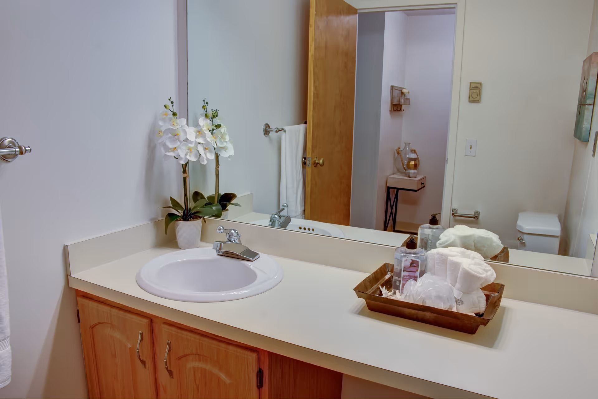 A bathroom countertop with a white sink, a wooden cabinet underneath, a large mirror above, a potted white orchid plant, and a tray holding rolled white towels and toiletries. A wooden door is partially open, revealing a toilet and a small table with a decorative item in the adjacent room.