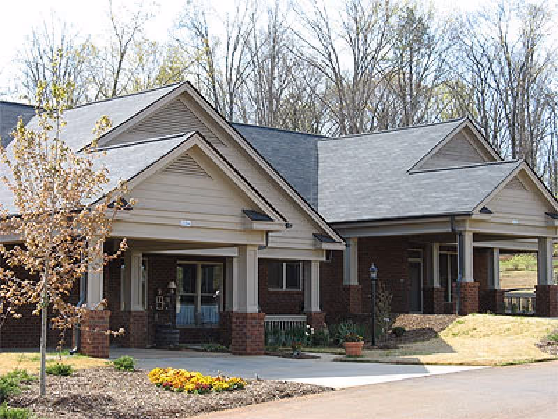 Exterior view of a single-story building with a gray roof and brick and beige siding, surrounded by a landscaped area with small trees and flowers under a clear sky.