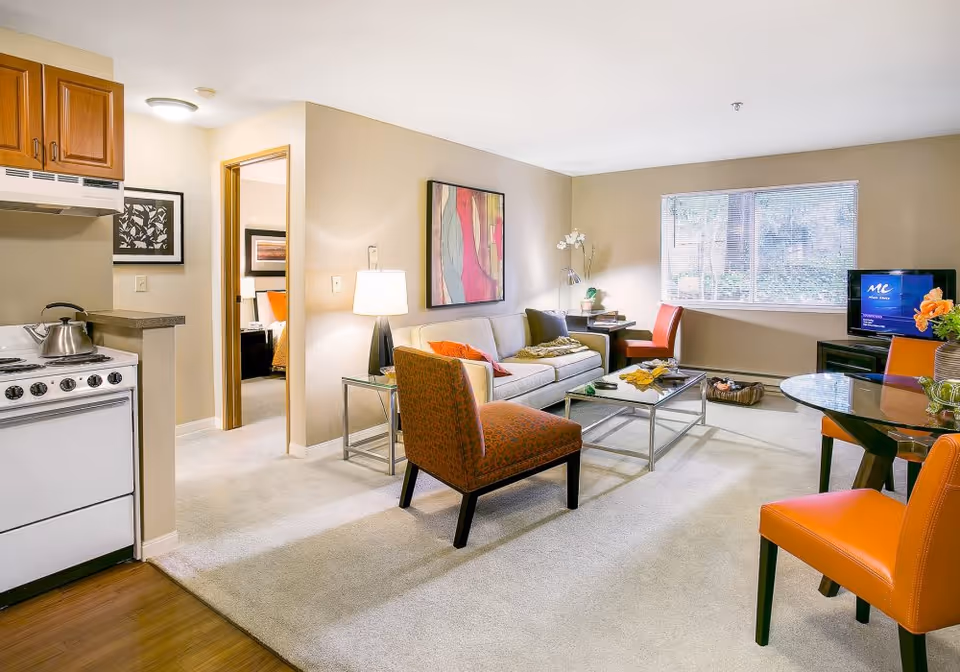 A bright and cozy living area in a senior living facility featuring a beige sofa with colorful pillows, an orange patterned armchair, a glass coffee table, and a round glass dining table with orange chairs. The room has a large window with blinds, a TV on a stand, and a small kitchen area with a stove and wooden cabinets. A doorway leads to a bedroom with a visible bed and artwork on the walls.