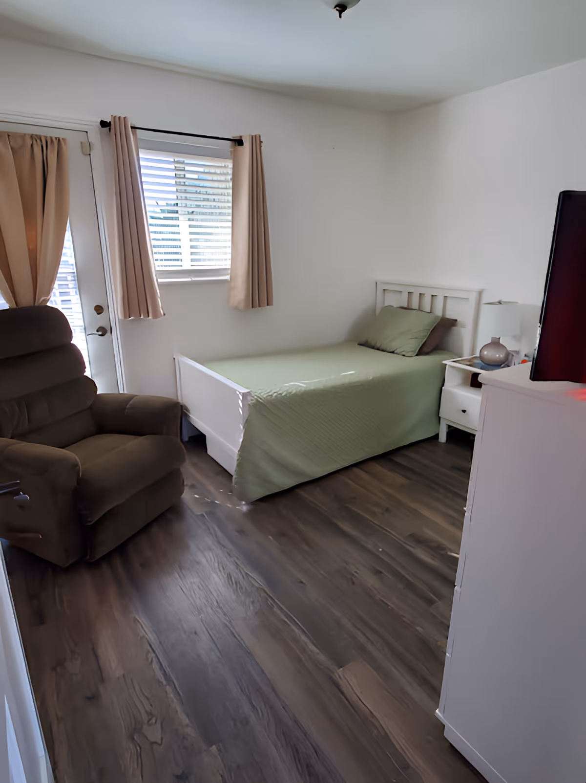 Small, simply furnished bedroom with a single bed covered in green bedding, a recliner chair, nightstand and dresser on wood-look flooring under a window and curtained door.