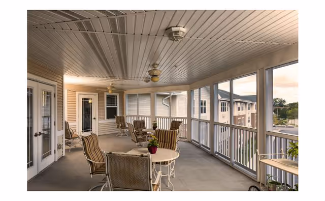 A spacious screened-in porch area with several cushioned chairs and small round tables, decorated with potted plants. The porch overlooks other buildings and greenery outside.