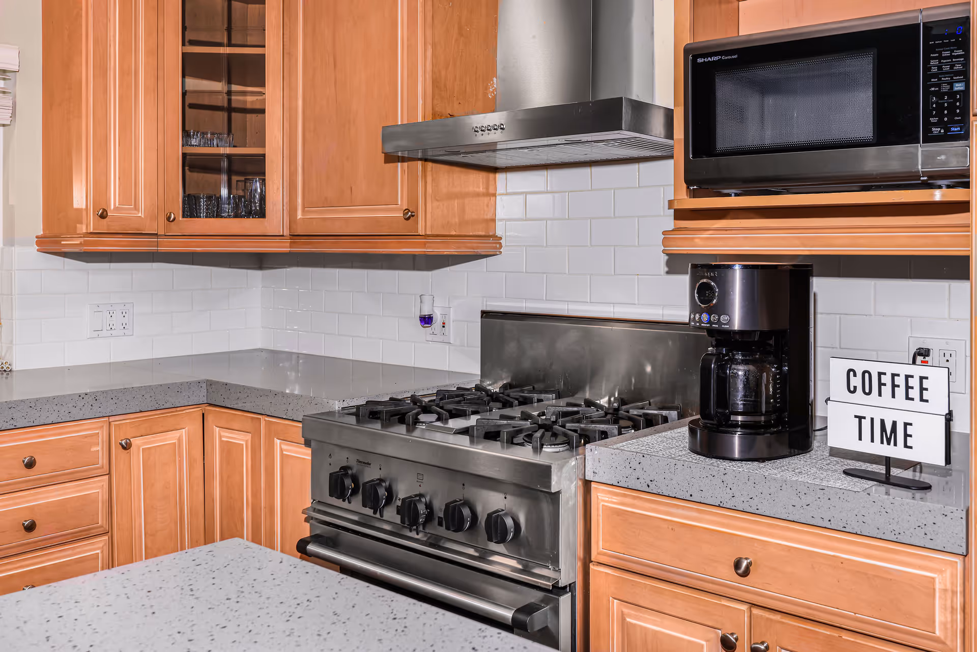 A modern kitchen with wooden cabinets, a stainless steel gas stove with an overhead vent, a microwave, a coffee maker, and a small sign that reads 'COFFEE TIME'. The countertops are gray with a speckled pattern, and the backsplash is white subway tile.