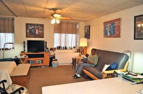 A cozy senior living room with TV, seating, lamps, and a resident reading.