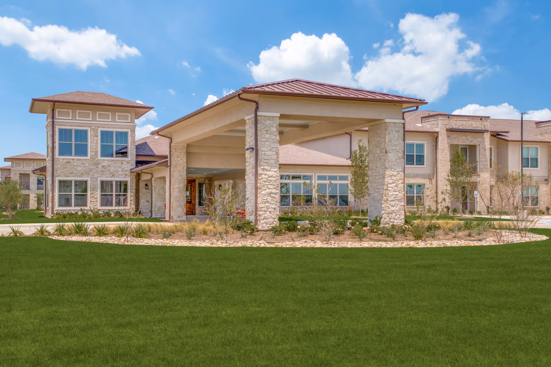Exterior view of Arabella of Red Oak Senior Living facility showing a large building with stone and beige walls, multiple windows, and a covered entrance supported by stone pillars. The foreground features a well-maintained green lawn and landscaped garden beds under a partly cloudy blue sky.