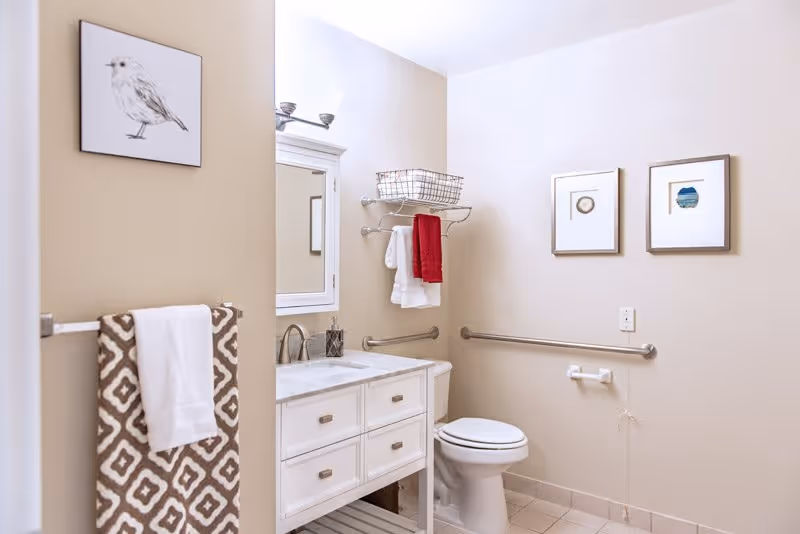 A clean and well-lit bathroom featuring a white vanity with a marble countertop, a mirror with a white frame, a toilet, and beige walls adorned with framed artwork. There are towels hanging on a rack and a shelf above the toilet with additional towels and a basket. Safety grab bars are installed near the toilet.