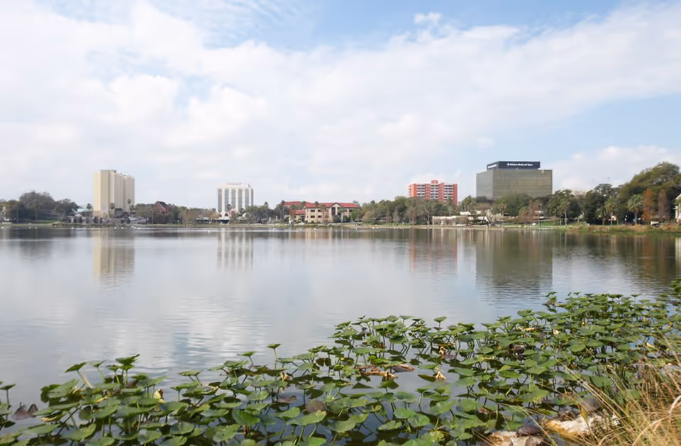 View across a calm lake with green lily pads in the foreground and a cityscape with various buildings in the background under a partly cloudy sky.