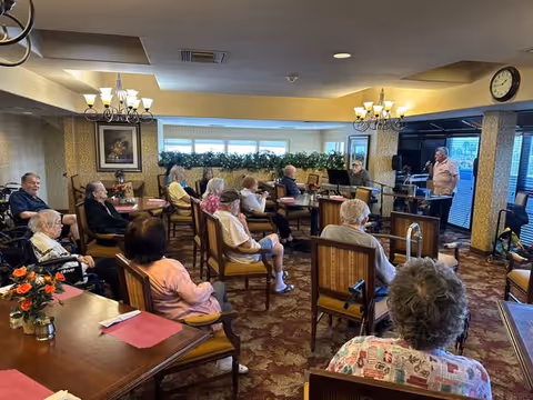 A group of elderly people seated in a common room of an assisted living facility, attentively listening to a man speaking at the front near a microphone and music stand. The room has warm lighting, chandeliers, floral carpet, and large windows with plants on the windowsill.