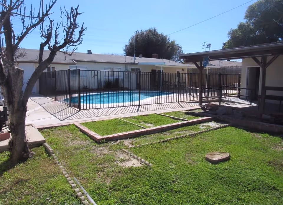 Outdoor area of a senior living facility featuring a fenced swimming pool surrounded by a concrete deck. There is a grassy lawn with a tree stump and a leafless tree in the foreground. A covered patio with wooden beams is visible on the right side, and single-story buildings are in the background under a clear blue sky.