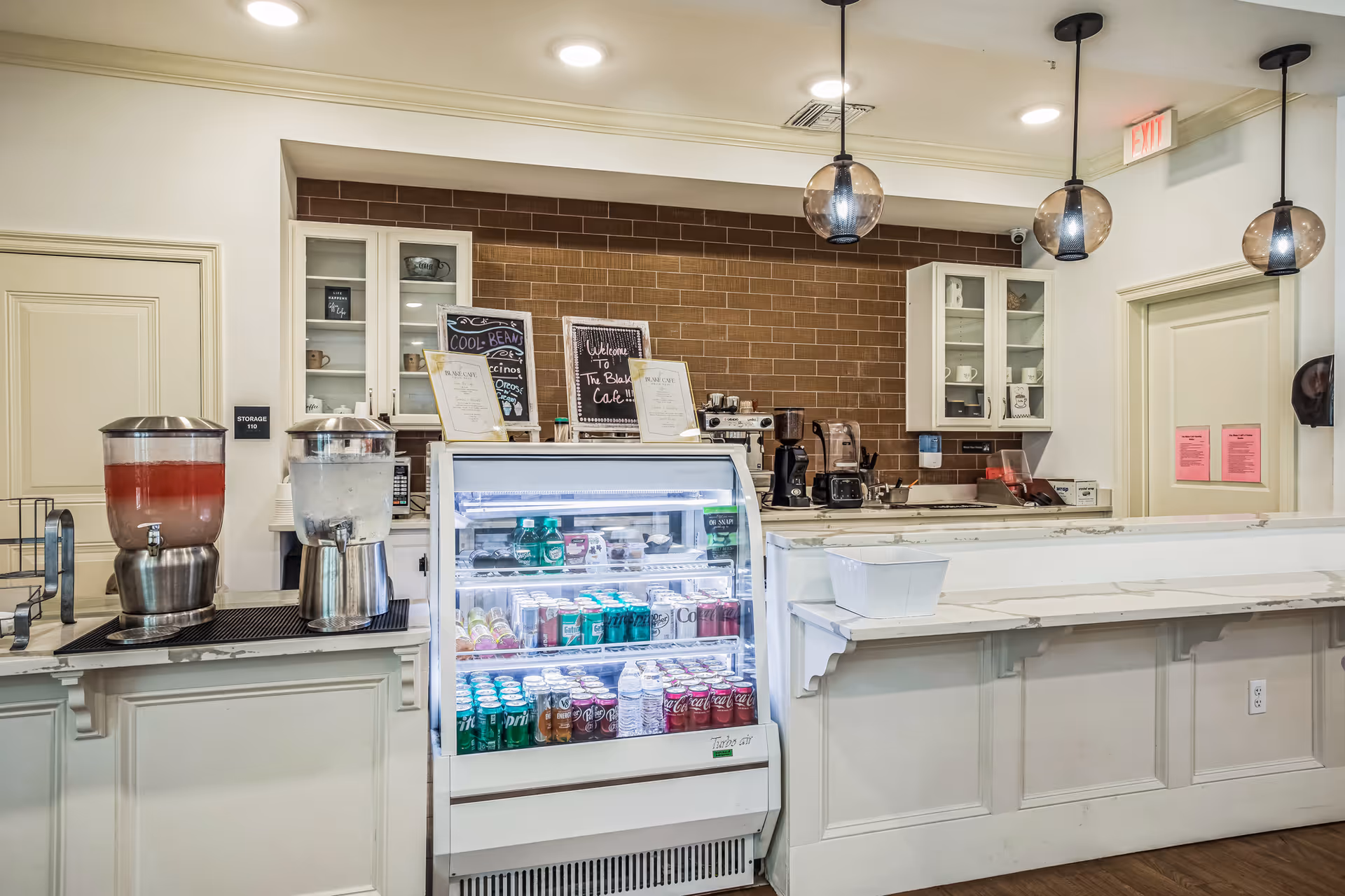 Interior view of a cafe counter area with a refrigerated display case filled with beverages, two beverage dispensers on the left, coffee machines and cups on the countertop, white cabinets with glass doors, and three hanging pendant lights above the counter.