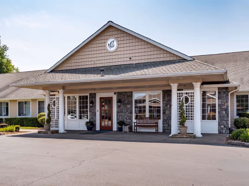 Front exterior view of McMinnville Memory Care facility showing a single-story building with a peaked roof, stone and beige siding, white columns, a red door, large windows, a wooden bench, and potted plants near the entrance.