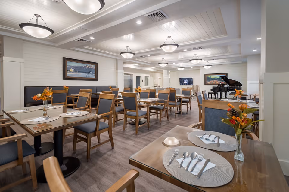 A spacious and well-lit dining room with multiple wooden tables and chairs arranged neatly. Each table has placemats, cutlery, and small vases with autumn-themed floral arrangements. The room features a light-colored ceiling with recessed lighting and hanging light fixtures. There are framed paintings on the walls, a flat-screen TV, and a grand piano in the background.