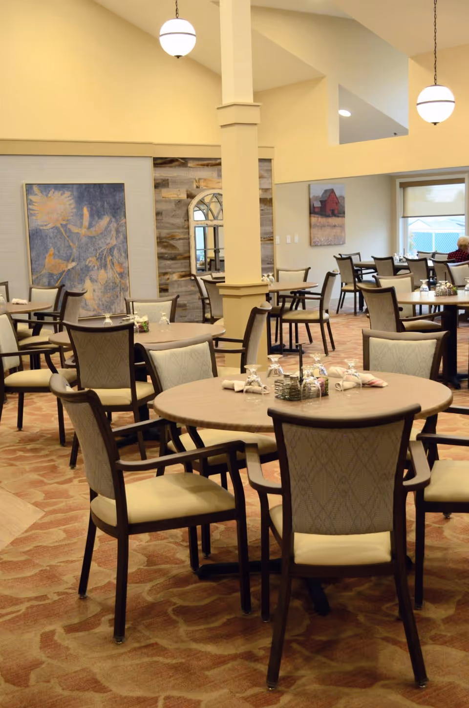 A spacious dining room with multiple round tables and cushioned chairs arranged neatly. Each table is set with upside-down glasses and folded napkins. The room features warm lighting with hanging globe lights, carpeted floors with a patterned design, and wall decorations including paintings and a mirror. A window with a roller shade is visible in the background.