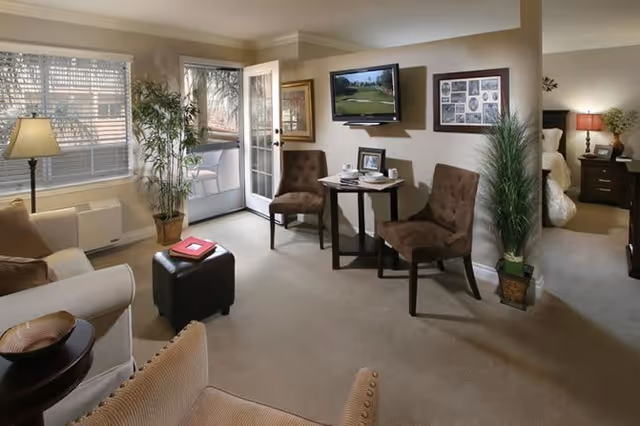 A cozy living room area with beige walls and carpet, featuring a beige sofa, two brown upholstered chairs around a small wooden table, a flat-screen TV mounted on the wall, a potted plant, and a door leading to an outdoor patio. A bedroom with a bed and nightstand with a lamp is visible through an open doorway.