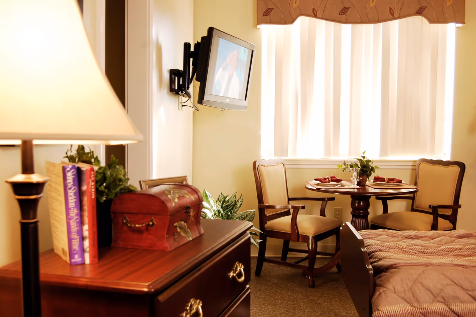 A cozy room with a wooden dresser holding a lamp, books, a decorative chest, and a plant. A wall-mounted TV is visible, and in the background, there is a small round table set with two chairs, napkins, and glassware near a window with vertical blinds and a valance. A bed with a brown quilt is partially visible in the foreground.