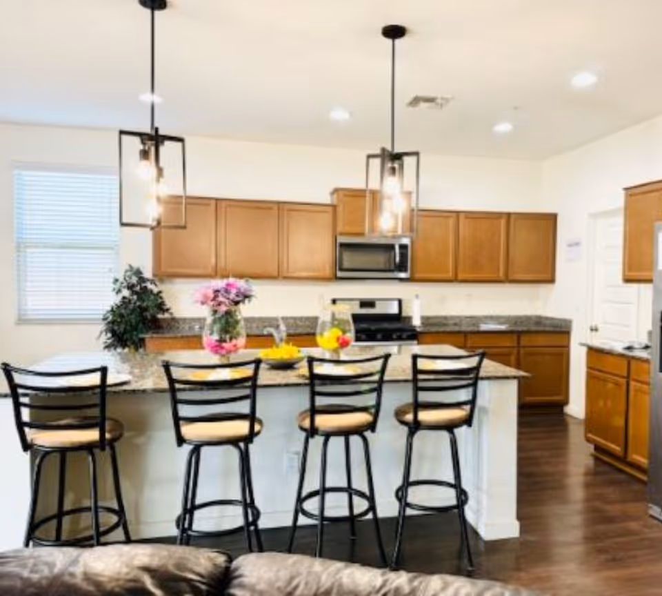A modern kitchen with wooden cabinets, granite countertops, and a kitchen island with four high chairs. The island has a vase with pink flowers and a bowl of fruit. Two pendant lights hang above the island. There is a stainless steel microwave and stove, a window with blinds, and a dark wood floor.