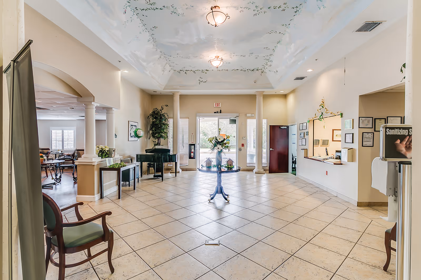 Bright assisted living lobby with tiled floor, a central table of flowers, piano, seating areas and a reception desk by the entrance.