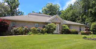 Single-story brick building with a triangular pediment above the entrance, surrounded by green bushes and trees, with a well-maintained lawn in the foreground under a clear blue sky.