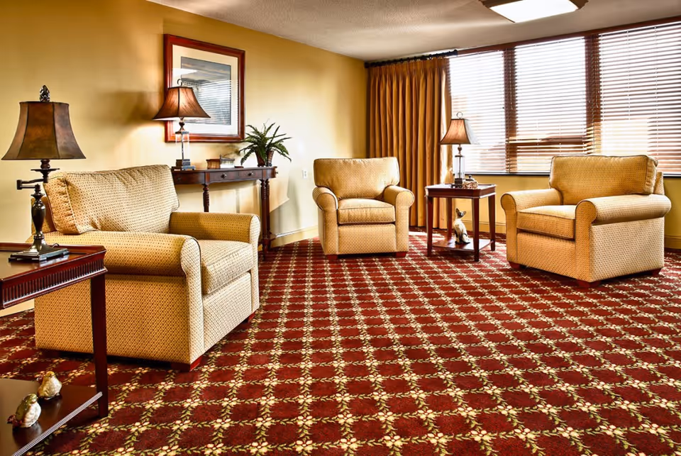 A cozy living room with three beige upholstered armchairs arranged around two wooden side tables with lamps. The room has a patterned red carpet, yellow walls, a framed picture on the wall, and large windows with wooden blinds and mustard-colored curtains.