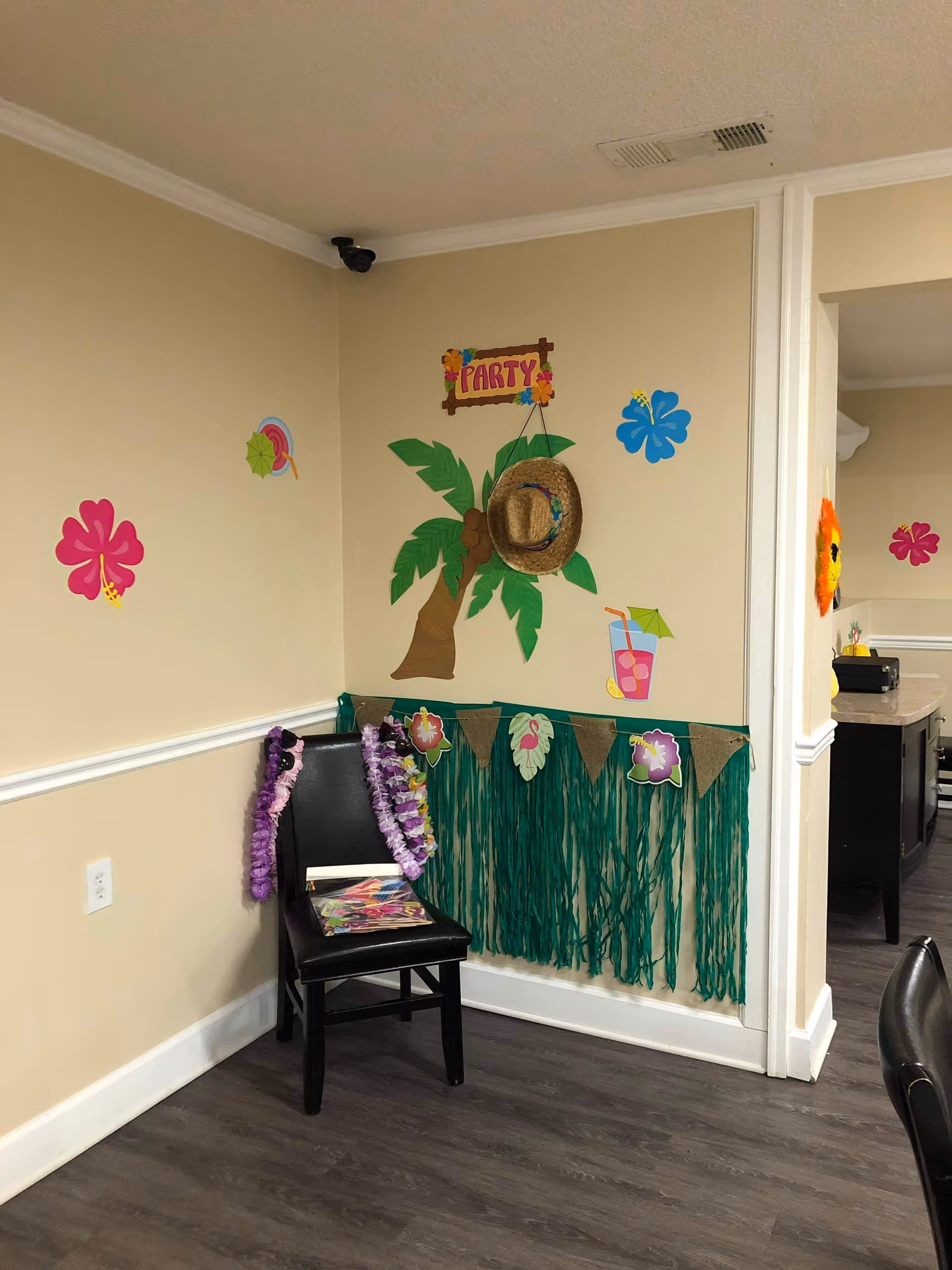 Corner of a room decorated with a tropical party theme, featuring a paper palm tree with a straw hat hanging on it, colorful paper flowers, a paper drink with a straw, and a green grass skirt decoration on the wall. A black chair with purple leis draped over it and some papers or magazines on the seat is placed against the wall. The room has beige walls, white trim, and dark wood flooring.