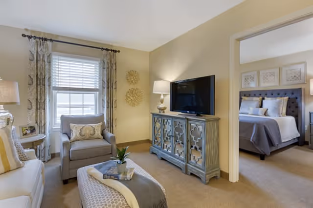A cozy living room with beige walls and carpet, featuring a gray armchair with a decorative pillow, a patterned ottoman with a throw blanket and a small plant, a TV on a decorative cabinet, and a lamp. Through an open doorway, a bedroom with a bed, pillows, and framed artwork on the wall is visible.