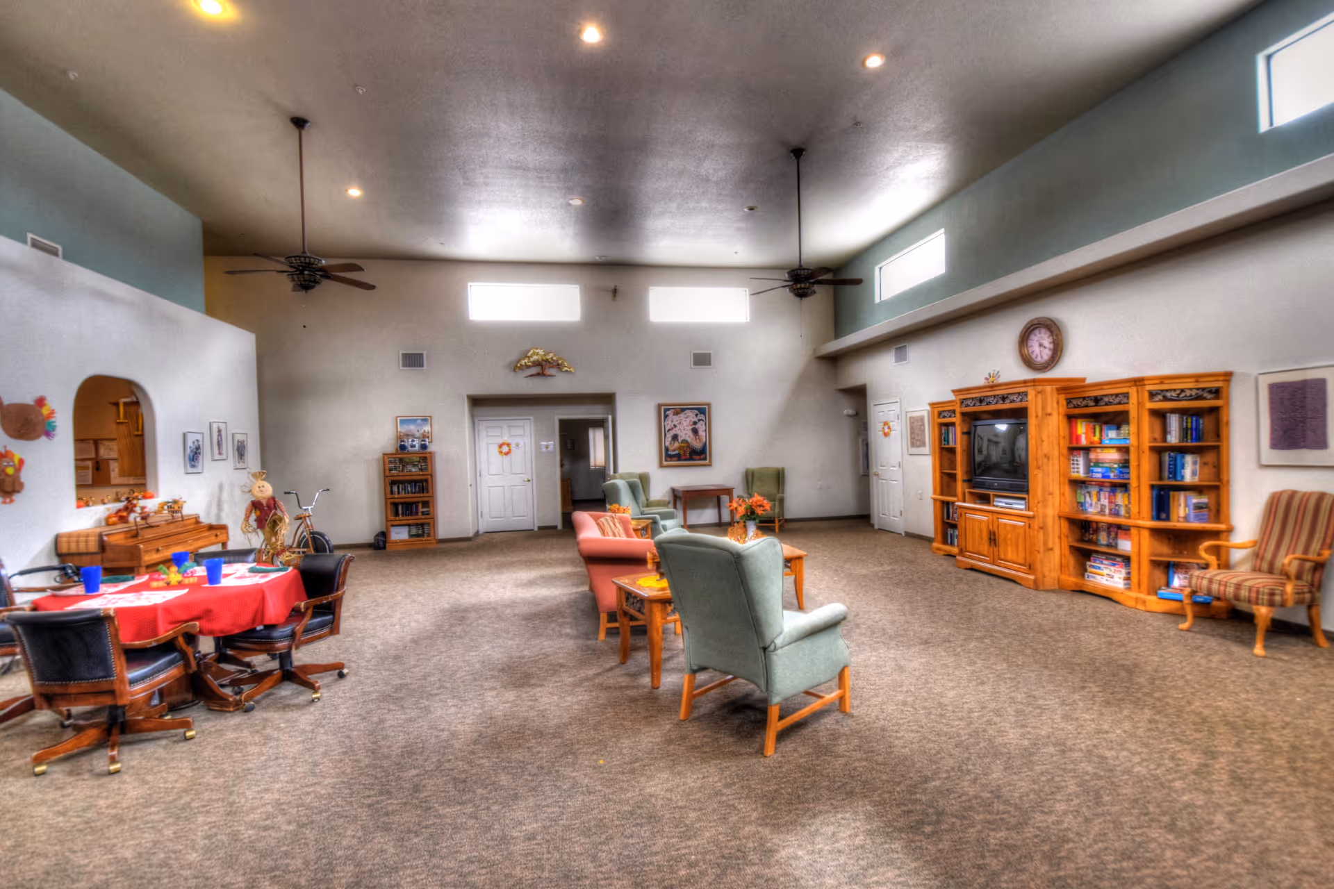 Spacious communal living area with armchairs, sofas, tables, bookshelves and a TV under high ceilings.