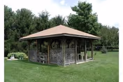 Wooden gazebo with lattice panels sitting on a grassy lawn surrounded by trees.