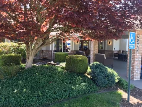 Outdoor view of a senior living facility entrance with a landscaped garden featuring a tree with red leaves, green bushes, and ground cover plants. There is a covered porch area with outdoor seating and a handicap parking sign in the foreground.
