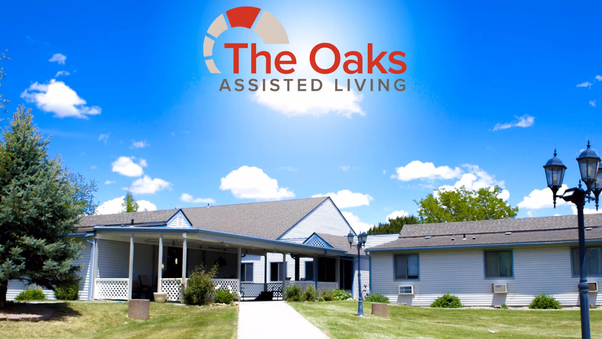 Exterior view of The Oaks Assisted Living facility showing a single-story building with a covered porch, surrounded by green grass, trees, and a clear blue sky with some clouds. There are two black lamp posts on the right side of the image.