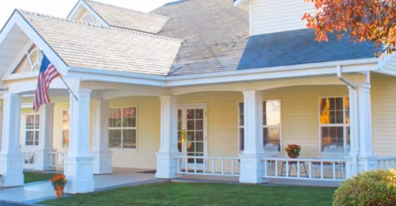 White-siding senior living facility with a covered front porch, columns, seating, and a flag.