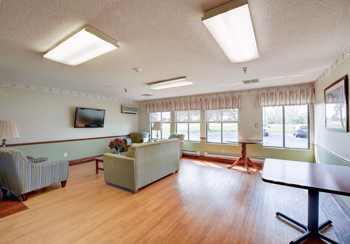 A bright and spacious living room in a senior care facility with large windows letting in natural light. The room features a green sofa, two armchairs, a coffee table with a vase of flowers, a wall-mounted TV, and two standing lamps. There are also two tables near the windows and a framed picture on the wall.