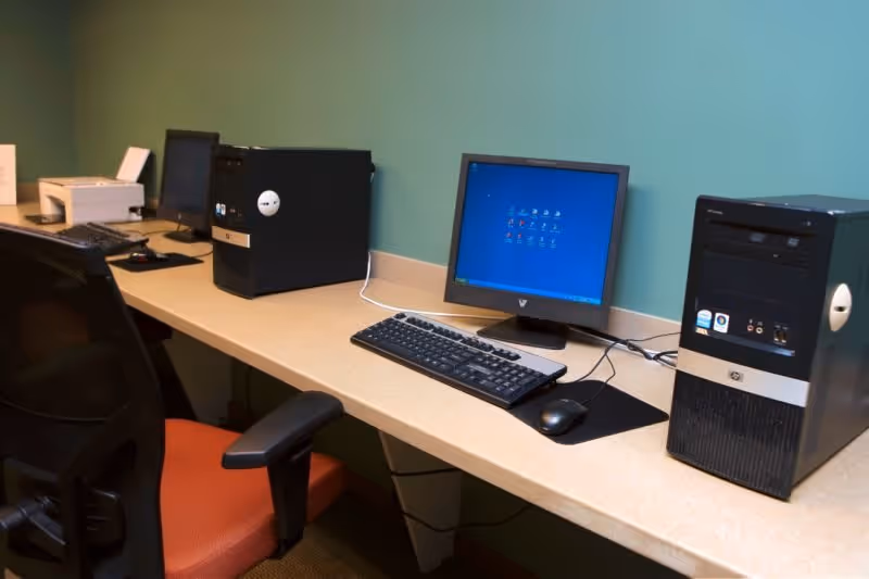 A computer workstation with two desktop computers, keyboards, mice, and a printer on a long beige desk against a teal wall. There is an office chair with a red seat in front of the desk.