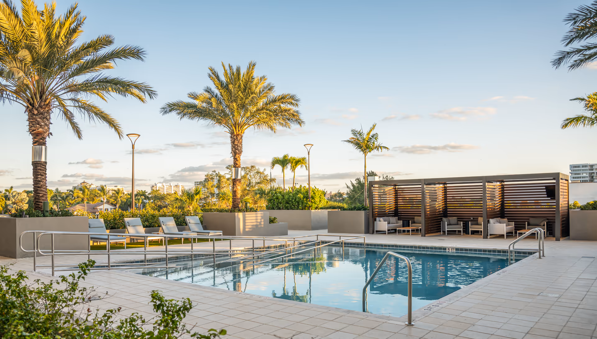 Outdoor swimming pool area with lounge chairs, palm trees, and shaded cabanas.