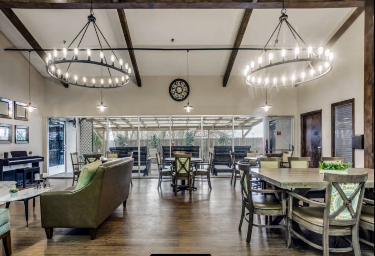 A spacious common area in a senior living facility featuring wooden floors, multiple tables and chairs, two large circular chandeliers hanging from a ceiling with wooden beams, a green sofa, a piano against the wall, and large windows with a view of an outdoor patio area with benches.