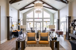 A spacious and well-lit living room with high vaulted ceilings and large windows allowing natural light to fill the space. The room features two patterned armchairs with orange cushions, side tables with decorative items, a coffee table, and additional seating areas with chairs and tables near the windows. The decor includes modern light fixtures, dark wood beams, and neutral-colored walls.